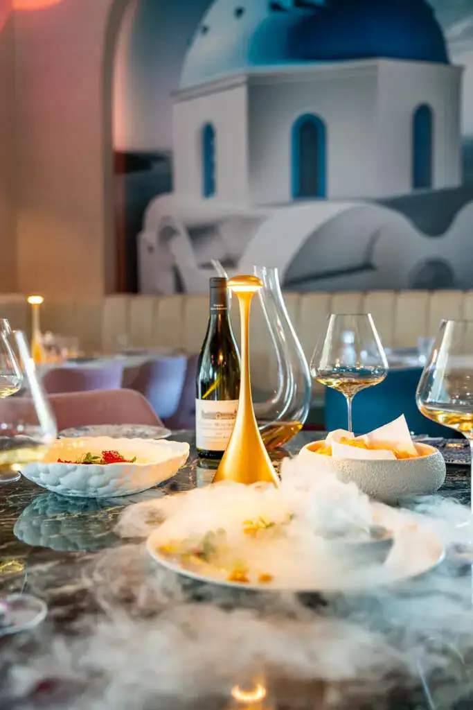 Elegantes Esstischarrangement mit Wein, Speisen und Stilleben in modernem, stilvollem Restaurant, inspiriert von der Architektur der griechischen Kirche Santorini.