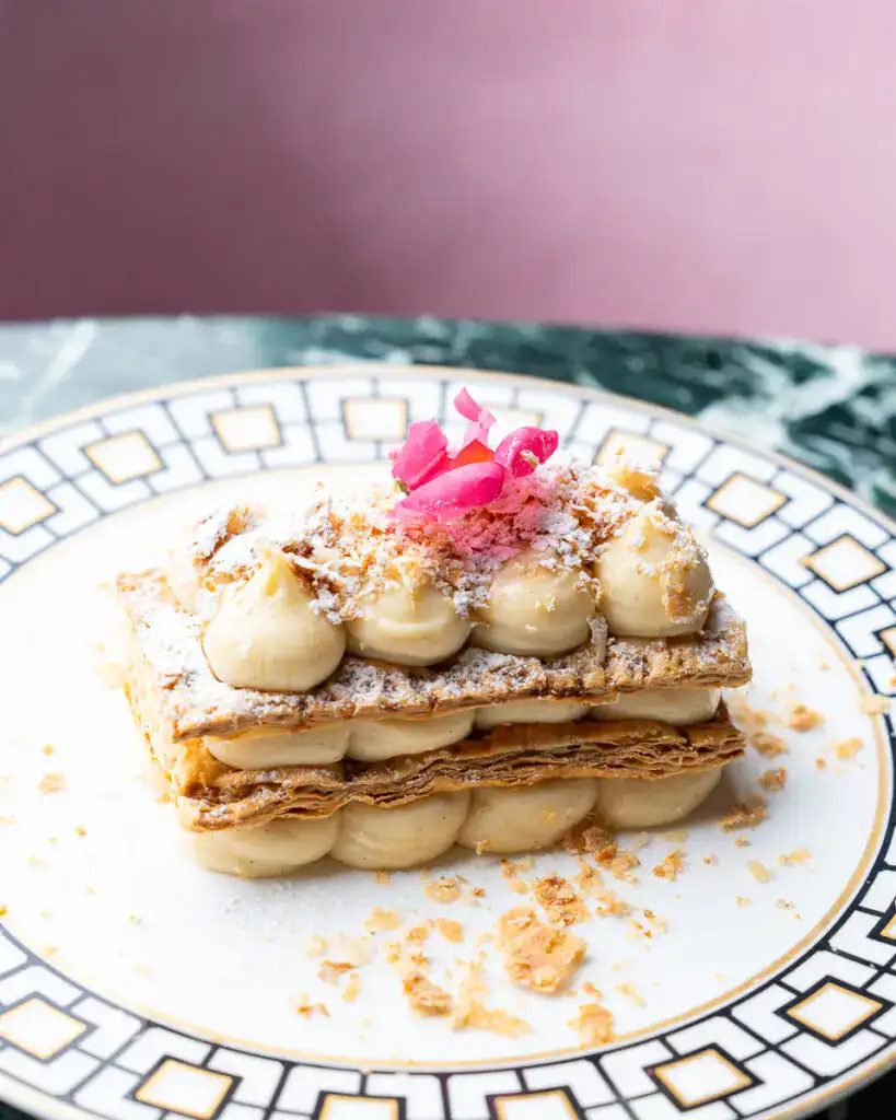 Kymata Modern: Elegante Dessert-Torte mit cremiger Vanillefüllung, knusprigem Blätterteig und zarten Rosenblüten, kunstvoll angerichtet auf einem stilvollen Teller. Perfekt für Gourmets und Dessertspezialisten.