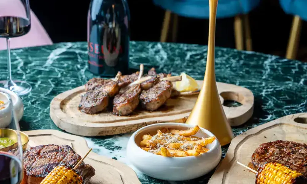 Gegrilltes Fleisch mit Maiskolben auf Holztellern, begleitet von Wein und cremiger Suppe, auf einem eleganten grünen Marmor-Tisch in einem modernen Restaurant.